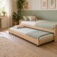 Cama nido infantil de madera clara con la segunda cama extraída, ropa de cama verde suave y una estantería baja junto a la ventana.